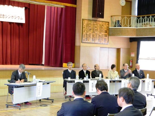 シンポジウムの会場の前方に司会者の席や、意見発表者の席が設けられており、右から二番目の男性がマイクを持って話をしている様子の写真